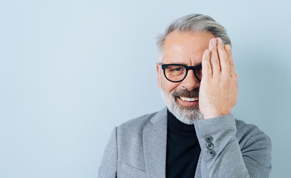 happy middle-aged man with eyeglasses looking at camera while covering his right eye with his hand